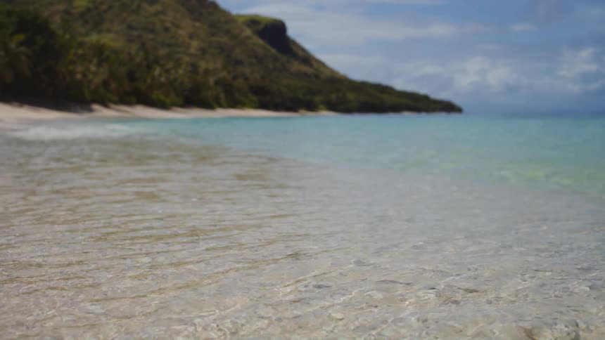 Gentle waves lap against a pristine white sand beach as the focus sharpens to reveal a lush tropical island coastline under a bright blue sky
