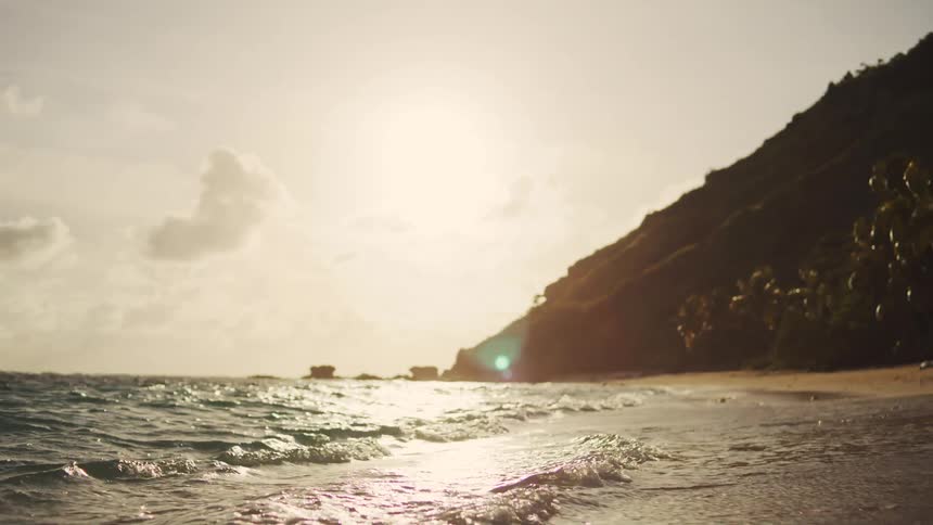 Gentle waves wash over the sandy shore as the sun glows brightly behind a silhouetted coastal cliff casting a golden shimmer across the water