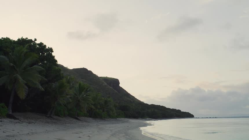 The camera pans slowly across a tranquil tropical beach at sunset showing soft waves lapping at the sand lush green palmcovered cliffs dominate the left side of the frame under a cloudy muted sky