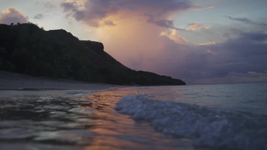 Gentle waves reach sand before forested cliff under colorful evening sky