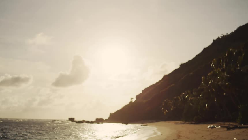 Bright sunlight illuminates a tropical beach where a lush green cliff meets the ocean