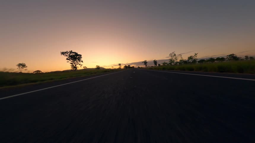 The video begins with a lowangle view driving down a twolane road towards a bright sunset with trees silhouetted along the horizon it then transitions to a side view flying over a grassy field as the road curves away