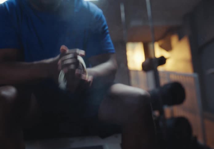 A person wearing a blue shirt and dark shorts sitting down vigorously rubbing white lifting chalk or powder between their hands he claps his hands together then stands up in a dimly lit gym