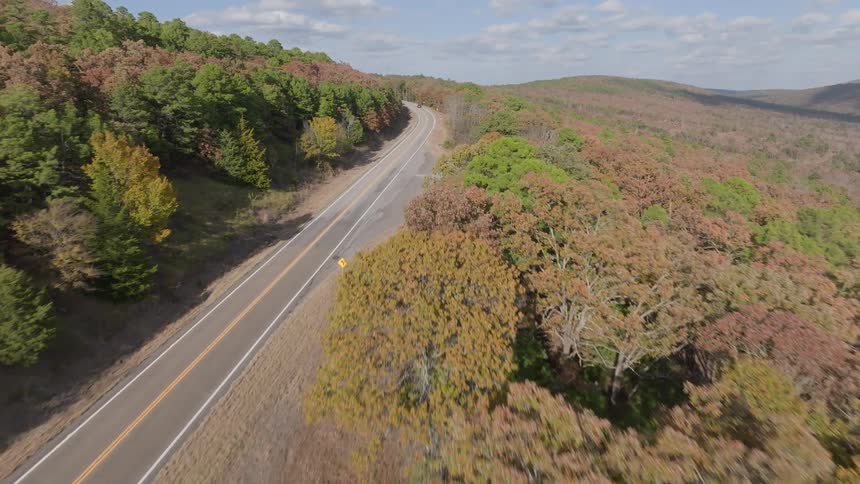 An elevated shot tracks forward along a winding twolane paved road that cuts through rolling hills covered in dense forest foliage showing a mix of green brown and autumnal colors as cars drive down the road
