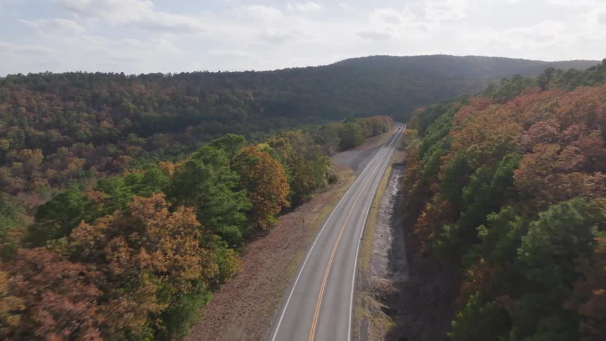 An aerial view shows a long straight road with vehicles traveling on is cutting through forested hills vibrant with autumn colors