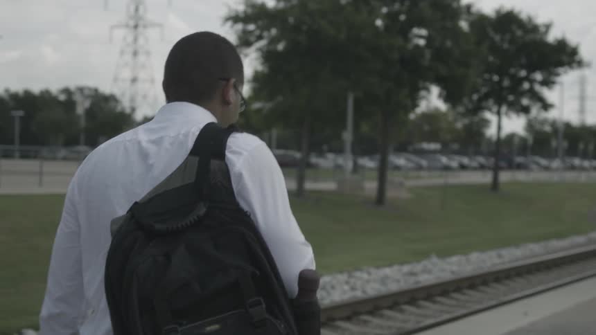 Man carrying a backpack on one shoulder walking next to some rails in a city on a cloudy day