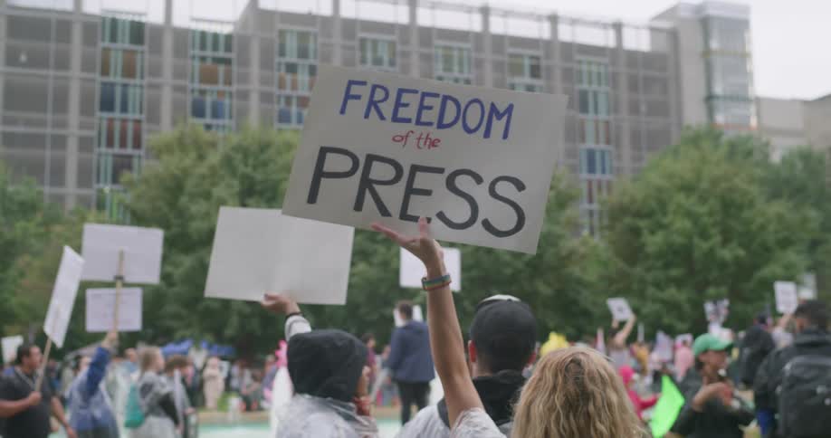 A closeup view focuses on a demonstrator holding a sign that reads freedom of the press above their head in a crowd the protest takes place outdoors in an urban setting with a large modern building visible in the background