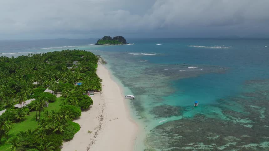 An aerial shot glides over a lush palm covered island with white sandy beaches and clear turquoise waters looking out toward a small rocky islet in the distance