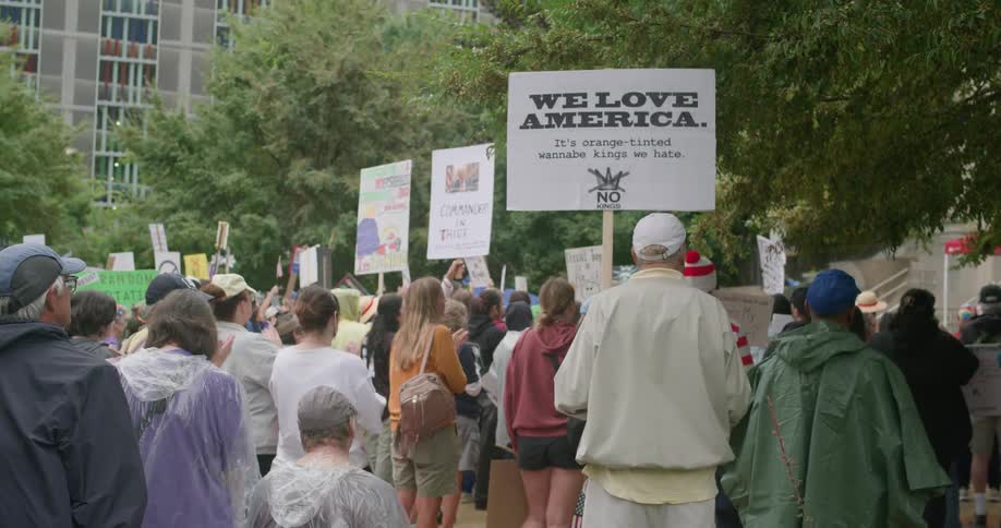 Person holds political message sign at outdoor protest surrounded by trees and costumes