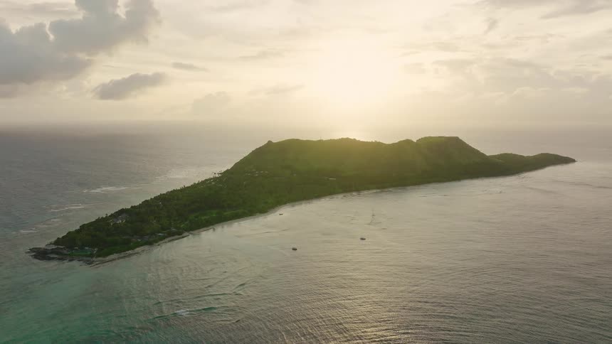 This is a high aerial drone shot of a narrow mountainous tropical island surrounded by the ocean the scene is captured during a sunset or sunrise with a bright hazy glow dominating the sky above the island illuminating the calm water around it