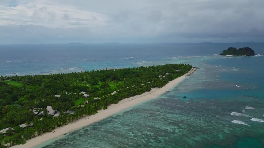 Tropical island with dense vegetation and scattered huts is surrounded by turquoise sea and visible reefs