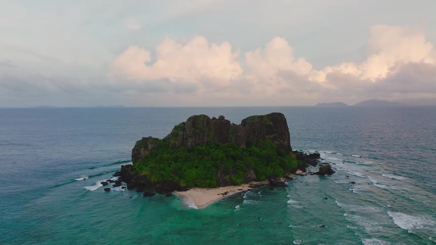 A mountainous island rises in the middle of the ocean at midday its slopes covered in lush green vegetation while waves crash against the rocky shoreline