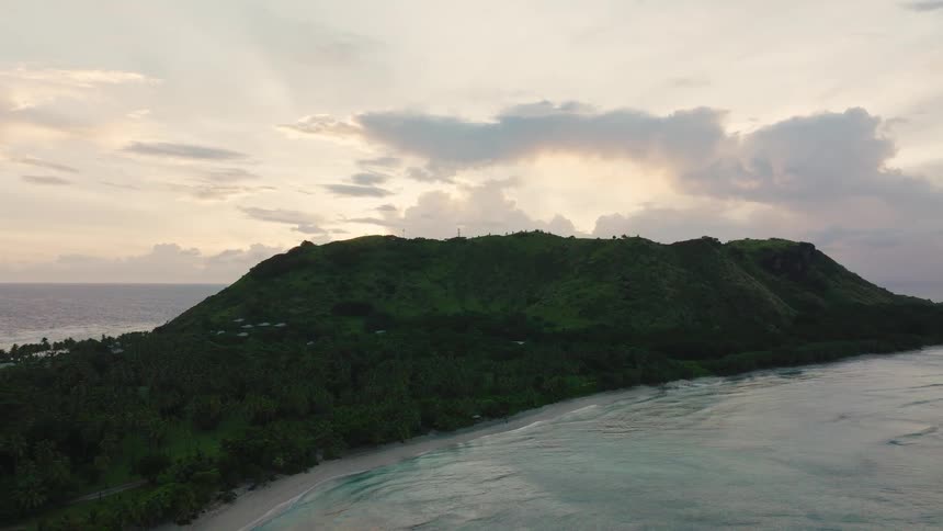 Hill covered in dense vegetation rises above tropical beach bathed by calm sea at sunset