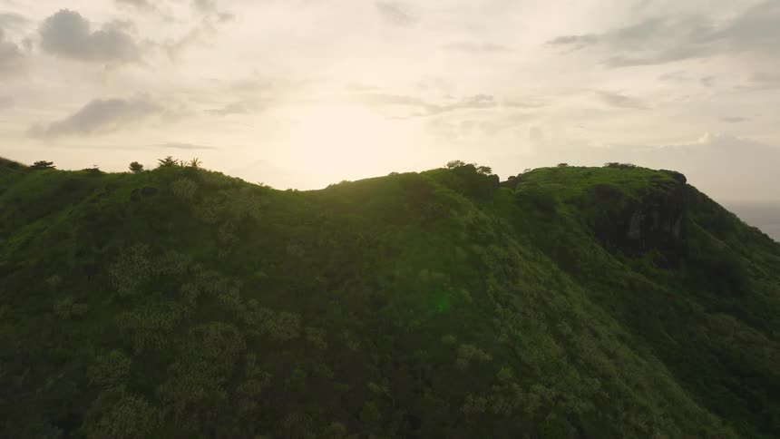 This is an aerial drone shot showing densely vegetated steep green mountains in a tropical or lush environment the camera slowly pans upwards and backward revealing a dramatic sunset or sunrise over the ocean as the brilliant light crests the ridge