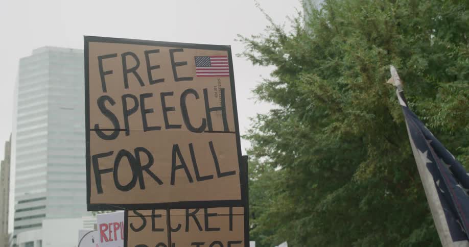 Sign with us flag advocates free speech at urban protest with buildings in background