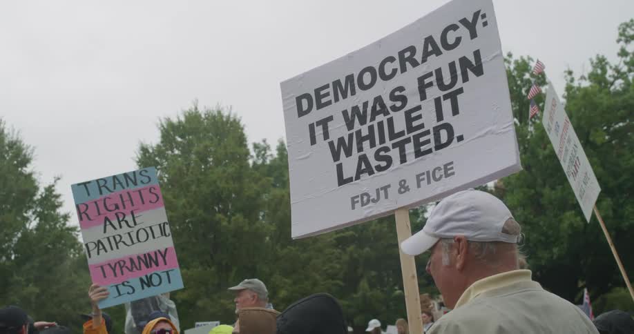 Sign with criticism of democracy is held by protesters among trees and security personnel