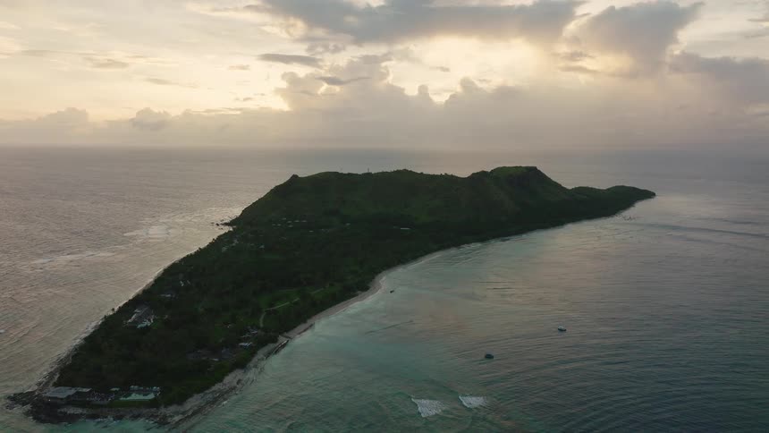An aerial shot captures a long narrow mountainous island covered in dark green foliage at sunset the island is surrounded by the deep blue ocean with turquoise water near the sandy shore
