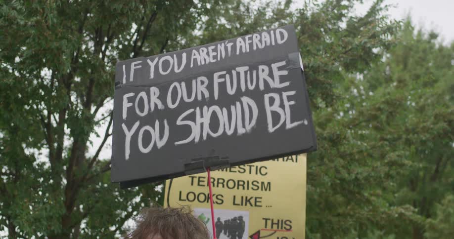 The prominent sign is black with white handpainted text that reads if you arent afraid for our future you should bea second partially visible yellow sign underneath has black text part of which reads not what domestic terrorism looks like