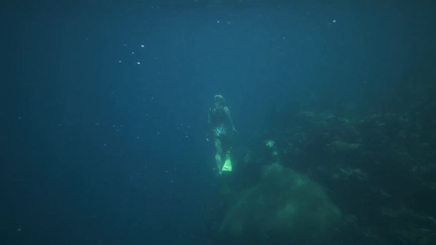 A freediver wearing yellow fins descends into deep blue water near a dark rocky seabed or reef the diver slowly approaches the bottom and turns to swim back toward the surface