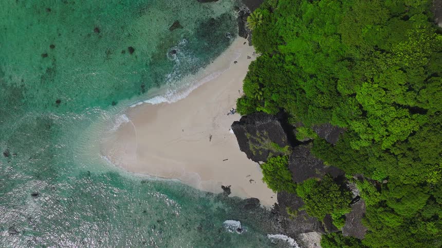 This is a high aerial shot looking down onto a pristine crescentshaped white sand beach nestled between steep dark rocky cliffs and a dense green forest the beach is bordered by shallow clear turquoise water