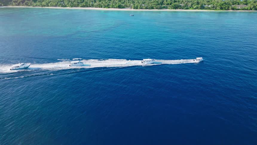 Four motorboats cut across deep blue waters past tropical island with lush greenery and pale beach
