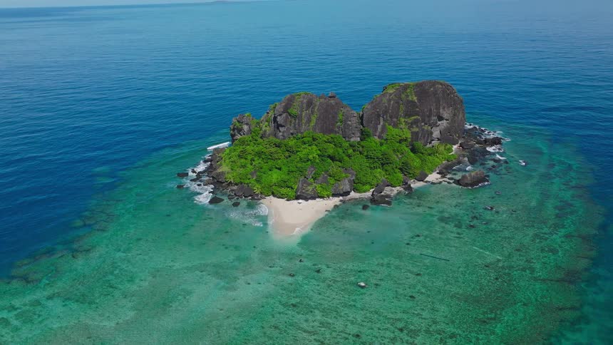 This video is an aerial view of a small lush green rocky island surrounded by vibrant turquoise and deep blue ocean waters a tiny patch of white sand beach is visible at the base of the steep twin rock formations