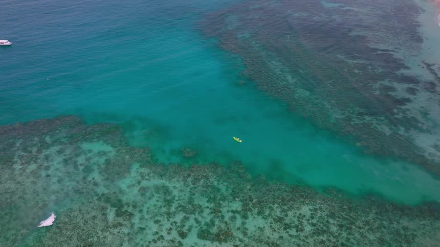 An expansive aerial view captures a vibrant turquoise ocean bordering a pinksand beach showing clear water over large dark coral reef formationsa small yellow kayak is seen paddling through a deeper channel between the reefs with two white boats a