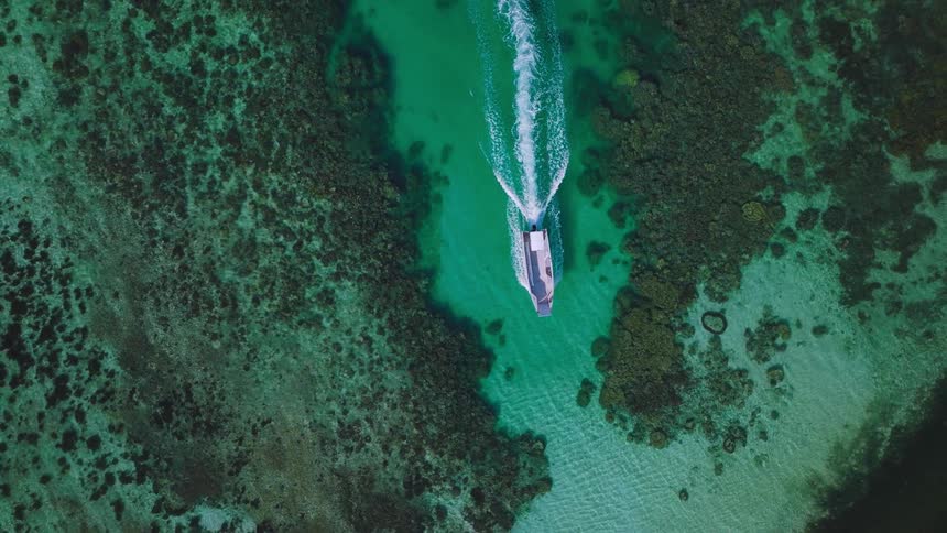An aerial view shows a speedboat moving across vibrant clear turquoise water leaving a white wake behind it the boat travels through a channel surrounded by dark green coral reefs and eventually approaches a sandy white beach
