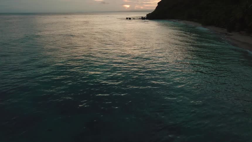 An aerial view captures dark ocean water at dusk with waves meeting a secluded sandy shore framed by a dark cliff
