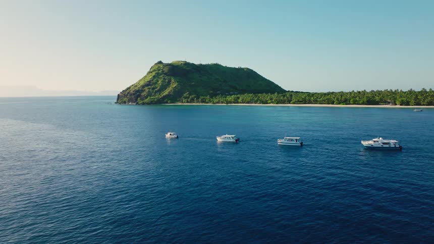 An aerial shot shows four small boats anchored in the deep blue ocean water near a tropical island the island features a large steep conical hill covered in lush green vegetation and a long palmfringed beach