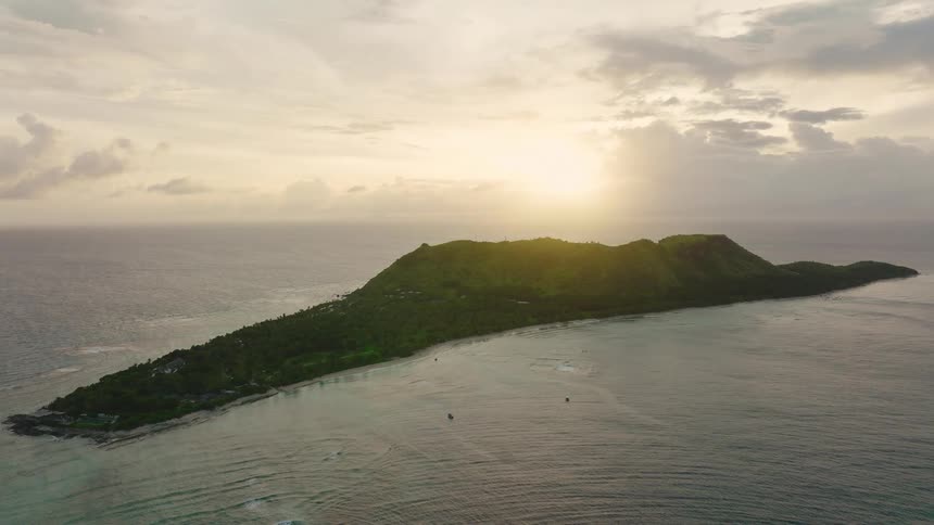 An elevated aerial shot captures a long narrow green tropical island with a distinct ridge line silhouetted against the bright hazy sunlight coming through the clouds over the ocean during sunset