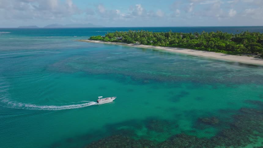 An aerial shot shows a small speeding motorboat carving a wake across vibrant turquoise tropical waters over a coral reef in the background is a lush green island with a white sand beach and a small rocky islet visible in the distance
