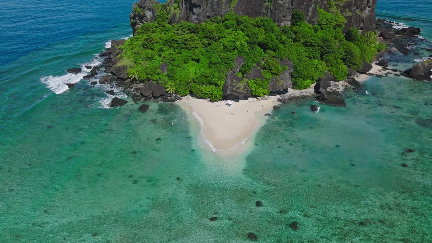 An overhead drone shot pulls back to reveal a small uninhabited tropical island featuring towering dark rock cliffs dense green vegetation on the peak and a tiny stretch of white sand beach where the clear turquoise water meets the shore