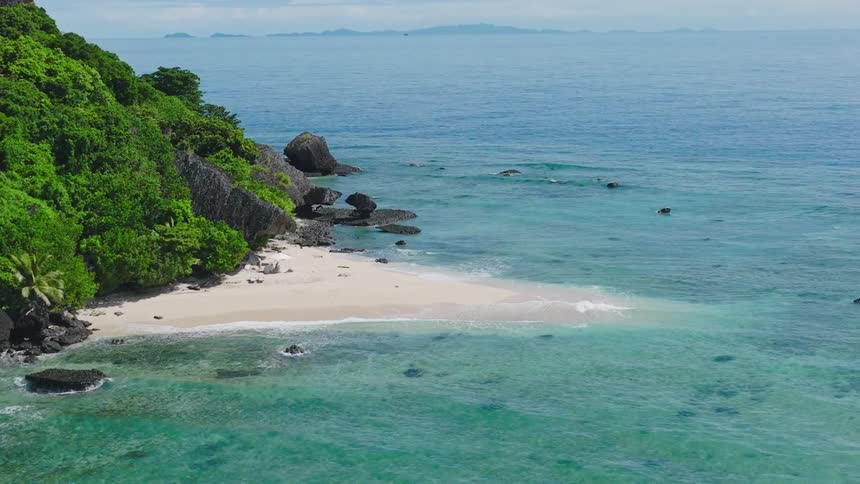Beach with pale sand and volcanic rocks borders blue ocean beside lush green cliffside
