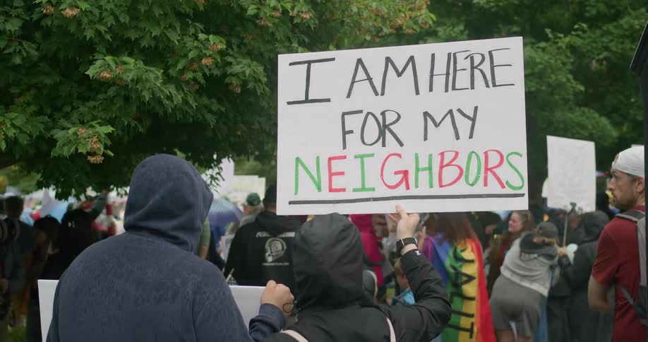Person holds sign reading i am here for my neighbors at rainy protest with rainbow flag in background