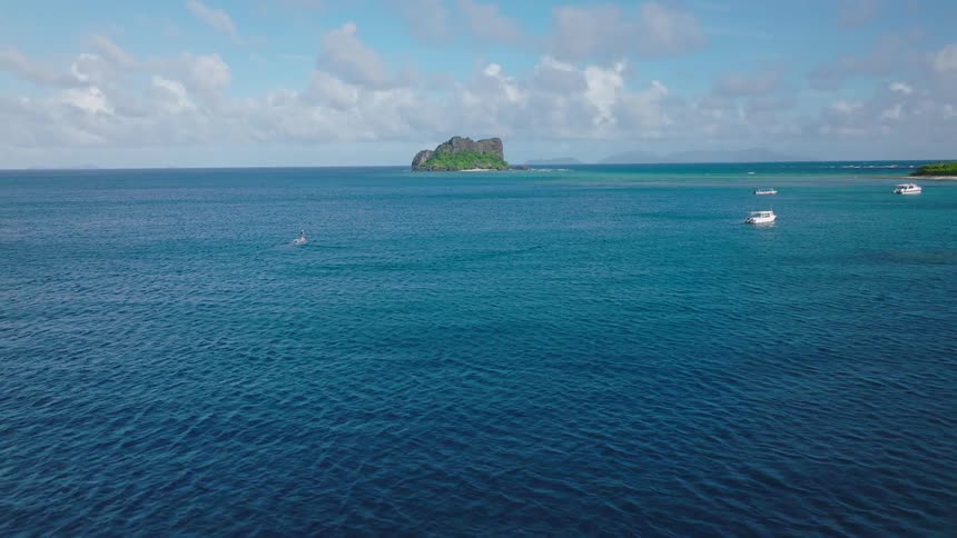 An elevated shot focuses on a person paddleboarding across the deep blue tropical ocean leaving a visible wake as they move toward the densely vegetated shoreline with a small rocky island visible further out in the water