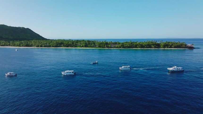 This aerial video captures a group of five small boats moving across the deep blue ocean near a lush tropical island with a prominent conical hill the clear bright sky and turquoise waters suggest a sunny day in a beautiful remote location