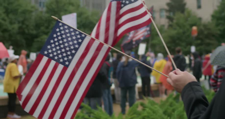 The video shows a protest or gathering where a person in the foreground is holding two small american flagsin the background a crowd of people is visible many holding signs suggesting a public demonstration and some appear to be wearing rain gear