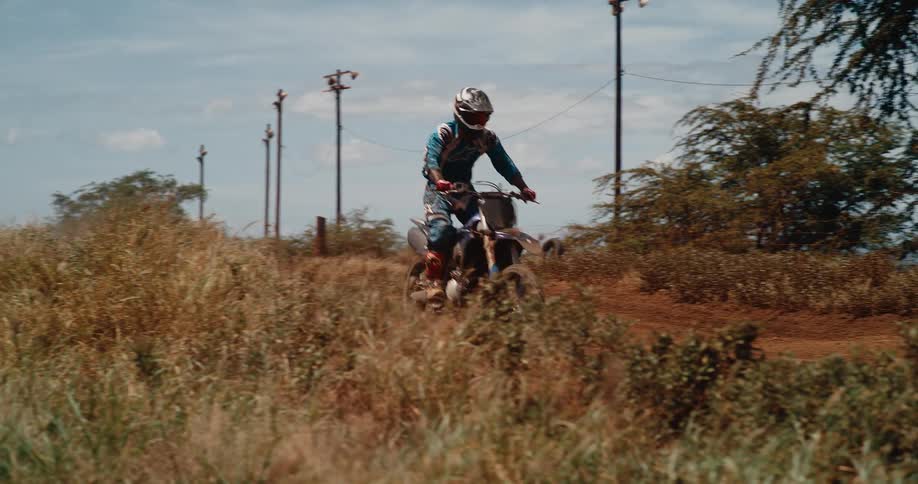 A dirt bike rider wearing full gear races over a grassy dirt track kicking up dust under a bright blue sky with power lines visible in the background