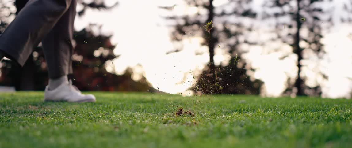 Club in motion approaches ball on short grass in treelined field