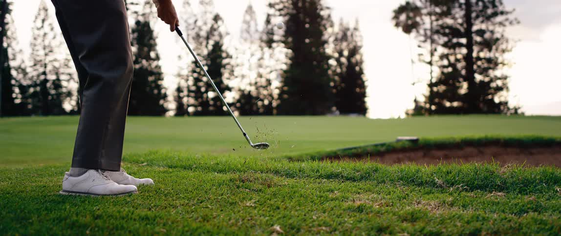 Club strikes ball over short grass near bunker on treelined course