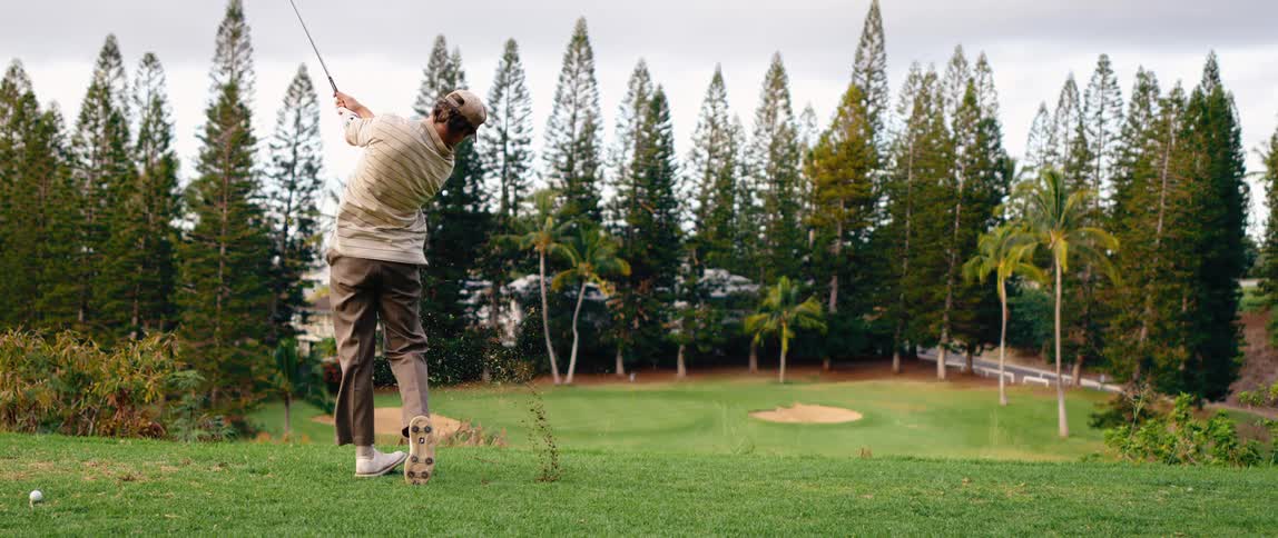 A man swings a golf club sending the ball and a large divot of grass flying from the green tee box surrounded by tall evergreen and palm trees