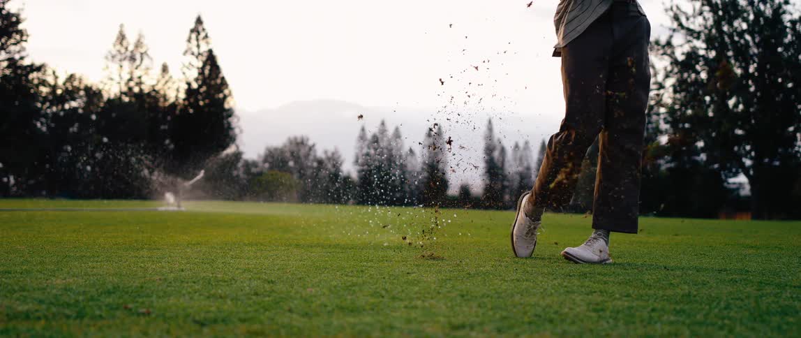 A man swings his club on a golf course at midday striking the ball off the grass as a sprinkler sprays in the background