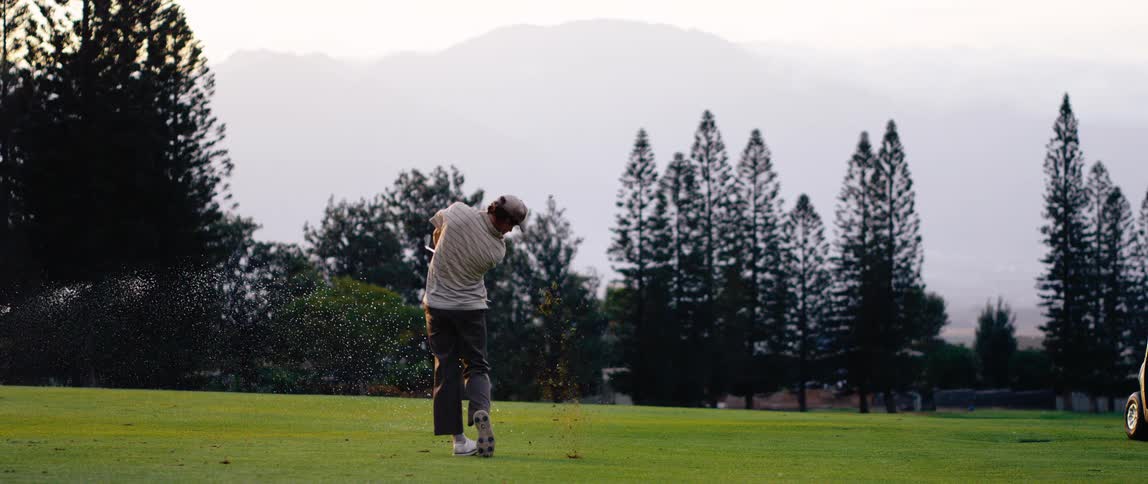 Golfer swings on green course surrounded by pines with misty mountains in the background