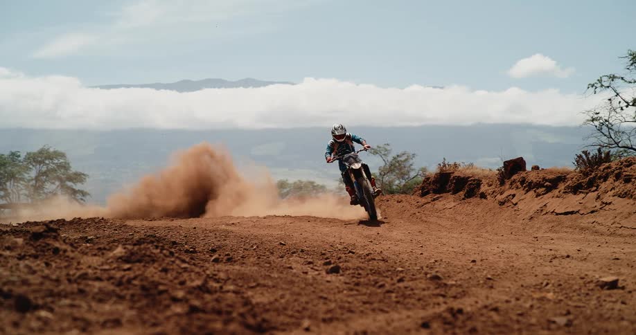 A dirt bike rider in full gear navigating a steep dusty turn on a red soil track leaving a large cloud of dust trailing behind the motorcycle with mountains and clouds visible in the distant background