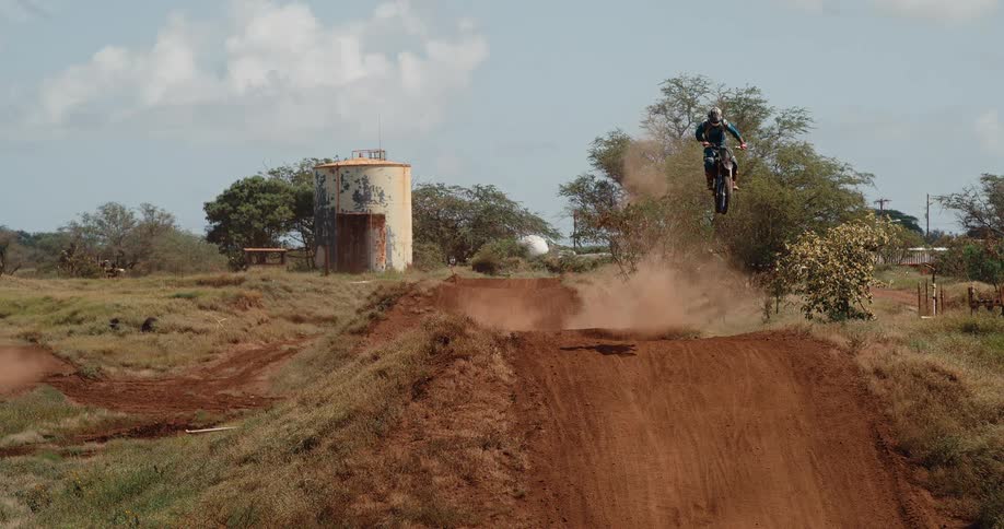 A dirt bike rider launches mid air over a dirt mound kicking up dust by scrubland