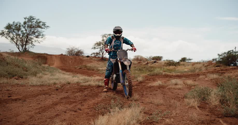 A wide shot shows a motocross rider fully geared up in blue and white racing attire sitting static on a dirt bike and looking forward centered on a reddishbrown dirt track with dirt ramps and dry vegetation surrounding the area under an overcast sky