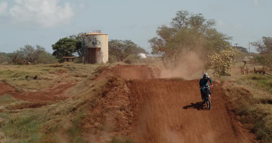 This video captures a motocross rider midjump on a dirt track showcasing a moment of action sportsthe rider launches over a large mound of red dirt kicking up a cloud of dust with a rusty water tower and dry scrubby terrain visible in the backgrou