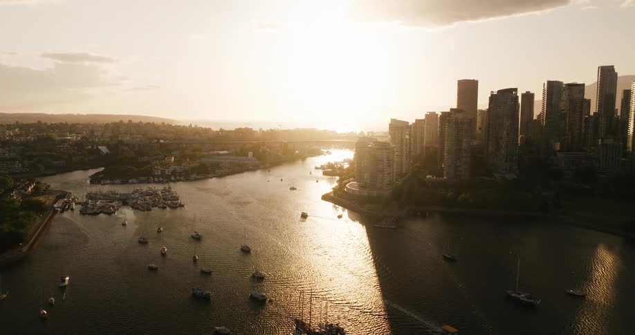An aerial view of a dense modern city skyline at sunset with tall buildings silhouetted against a bright sun over a body of water numerous boats and a marina are visible in the water which reflects the suns golden light