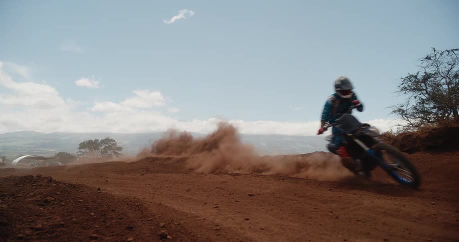 A motocross racer speeds along a dirt road at midday fully kitted leaving a trail of dust behind
